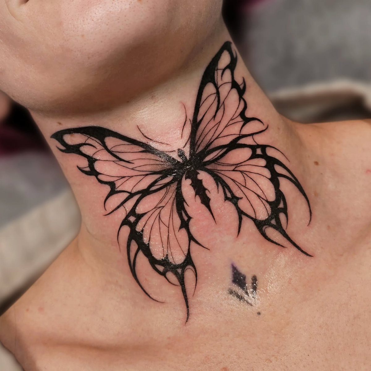 Stylized black butterfly tattoo centered on a person's neck with sharp wing details.
