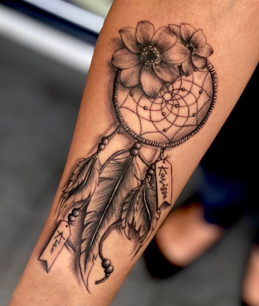 Dream catcher tattoo on a forearm with flowers, feathers, and name ribbons.