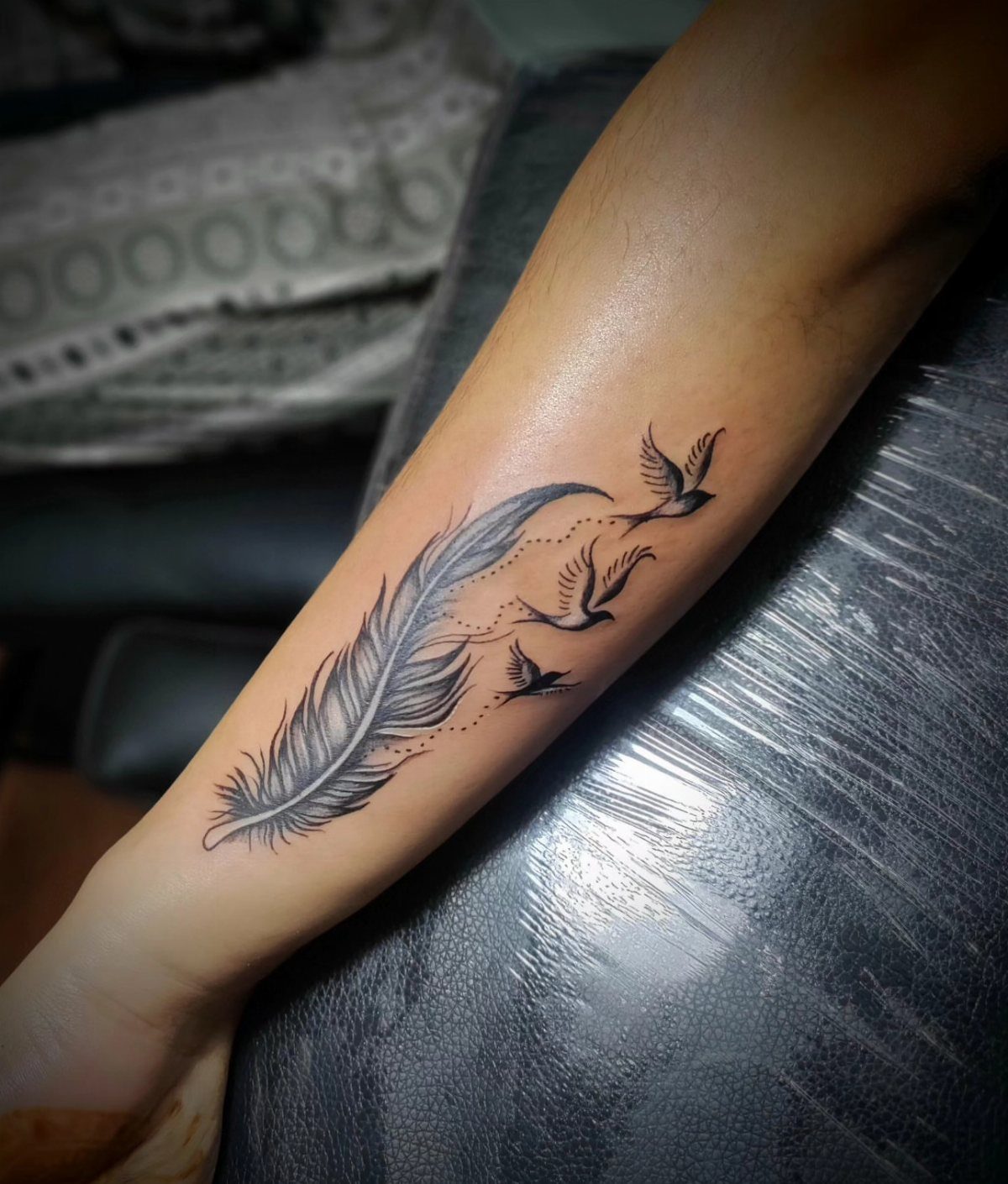 Feather tattoo on a forearm transitioning into three small flying birds in black ink