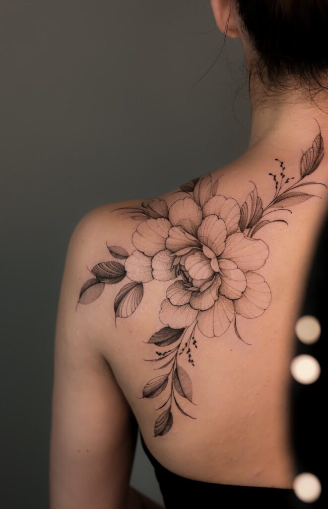 Large black ink peony flower tattoo on a person's upper back and shoulder.