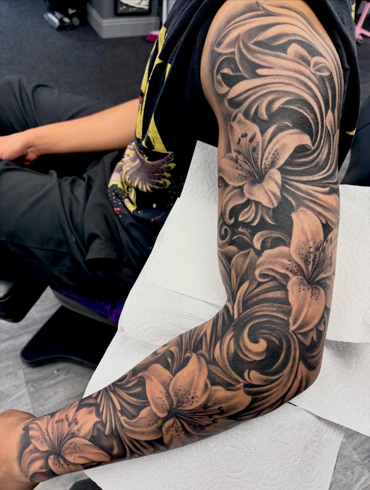 Full sleeve tattoo of lilies and swirls in black and grey ink on arm