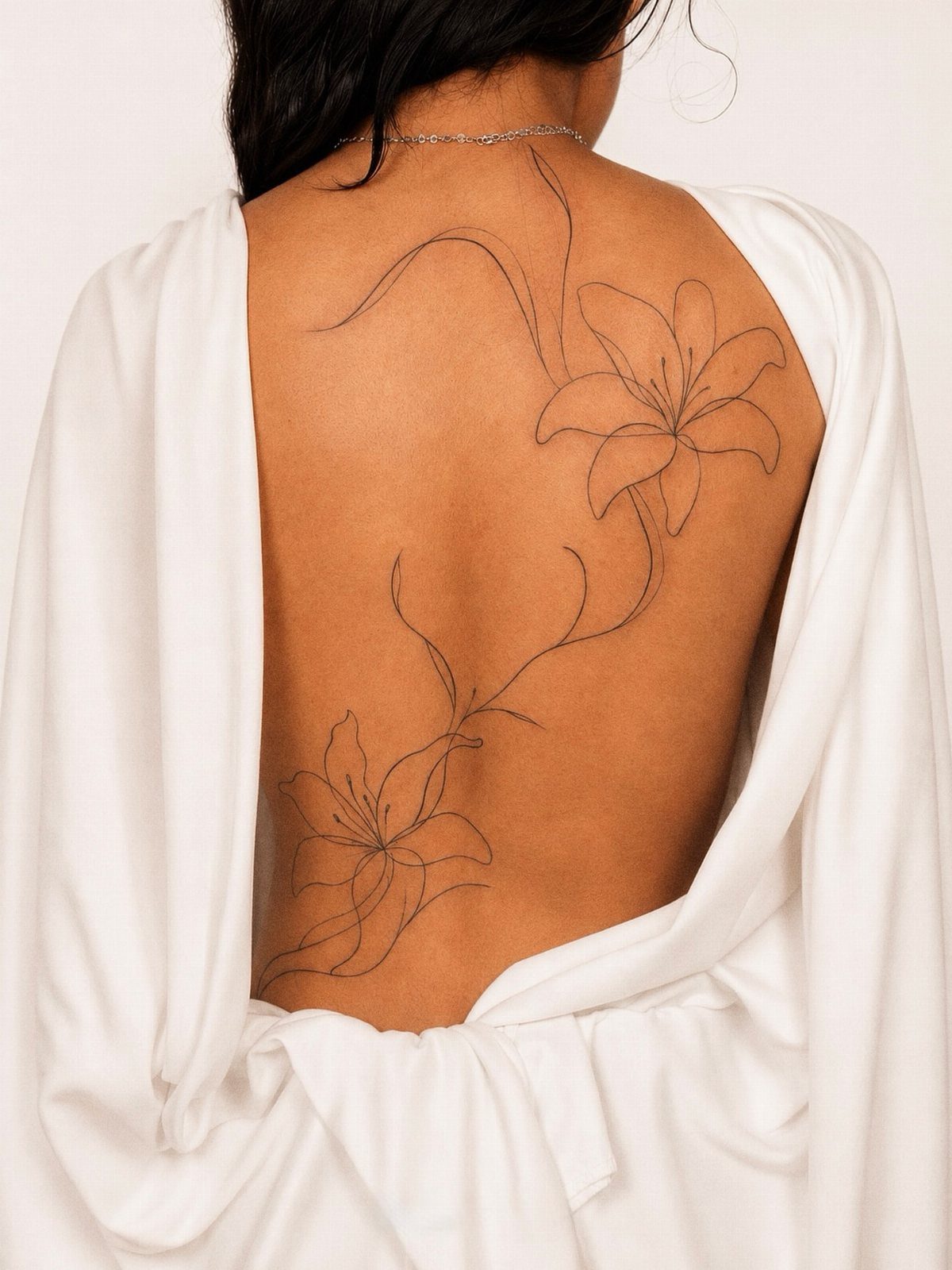 Fine-line lily flower tattoo stretching across the back of a person in white.