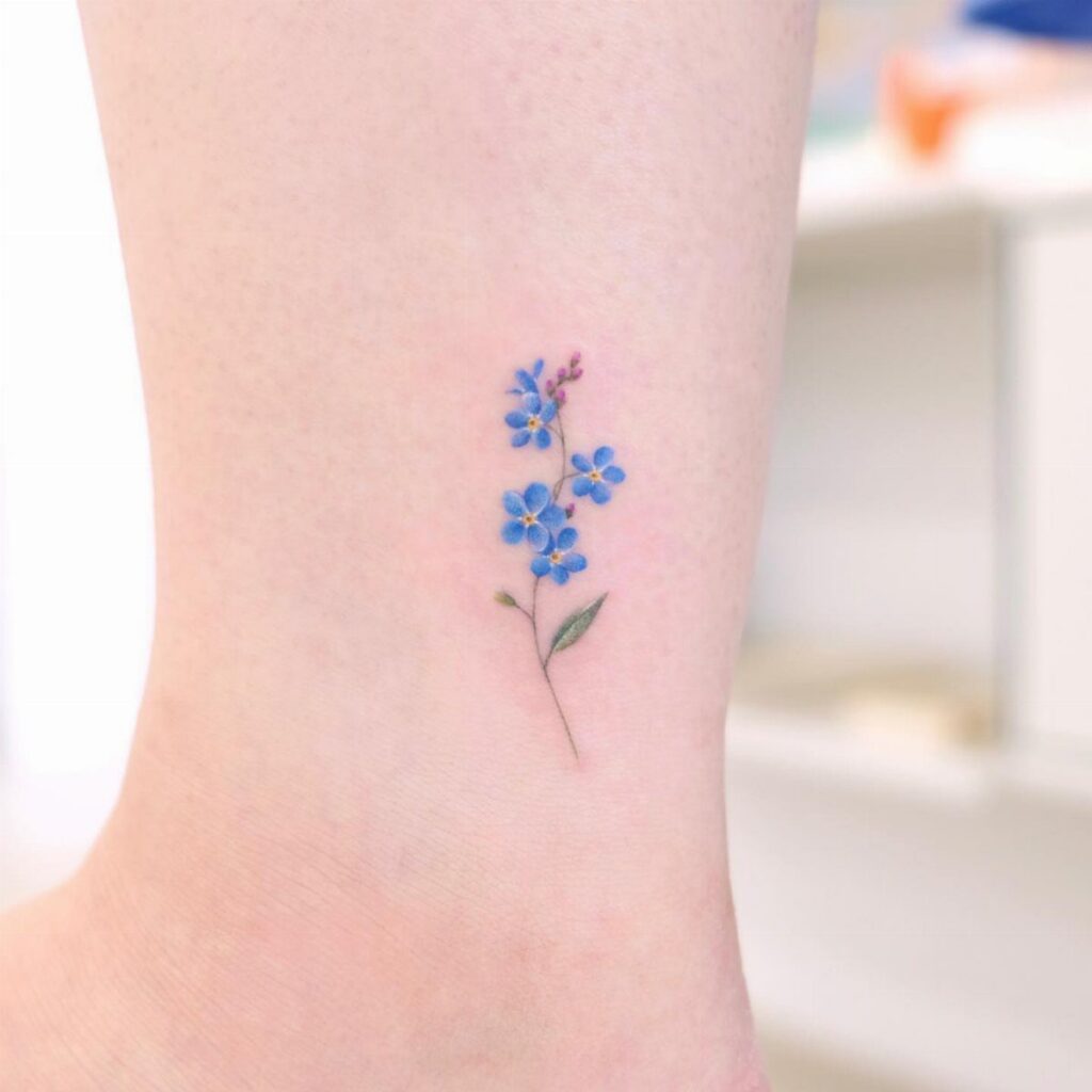 Small blue forget-me-not flower tattoo on a person's ankle with green stem.