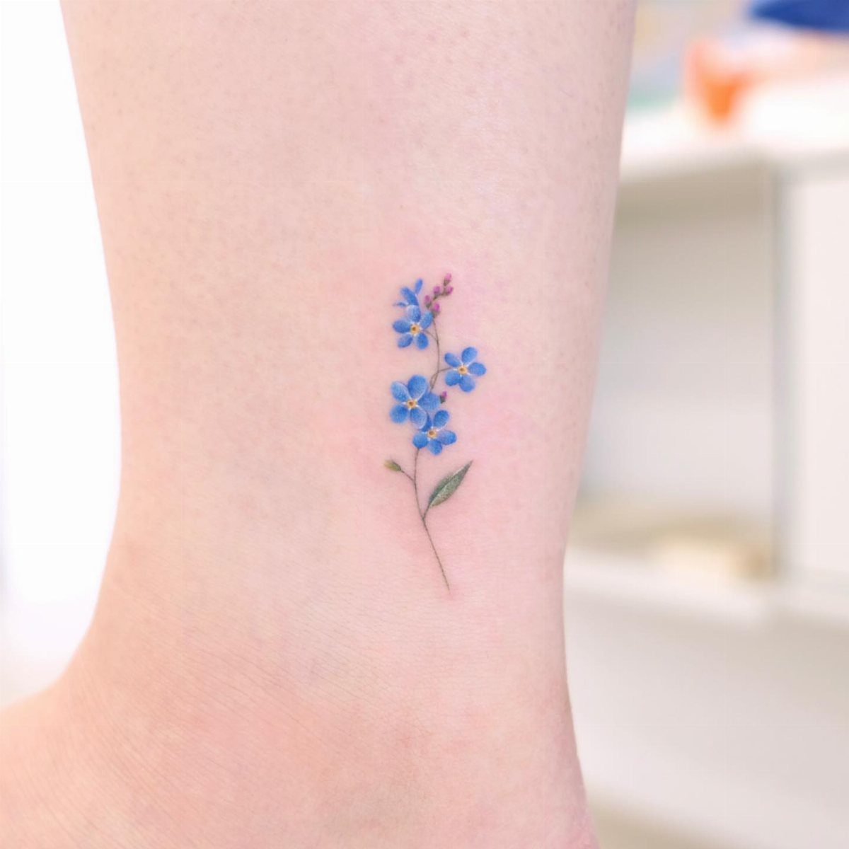 Small blue forget-me-not flower tattoo on a person's ankle with green stem.