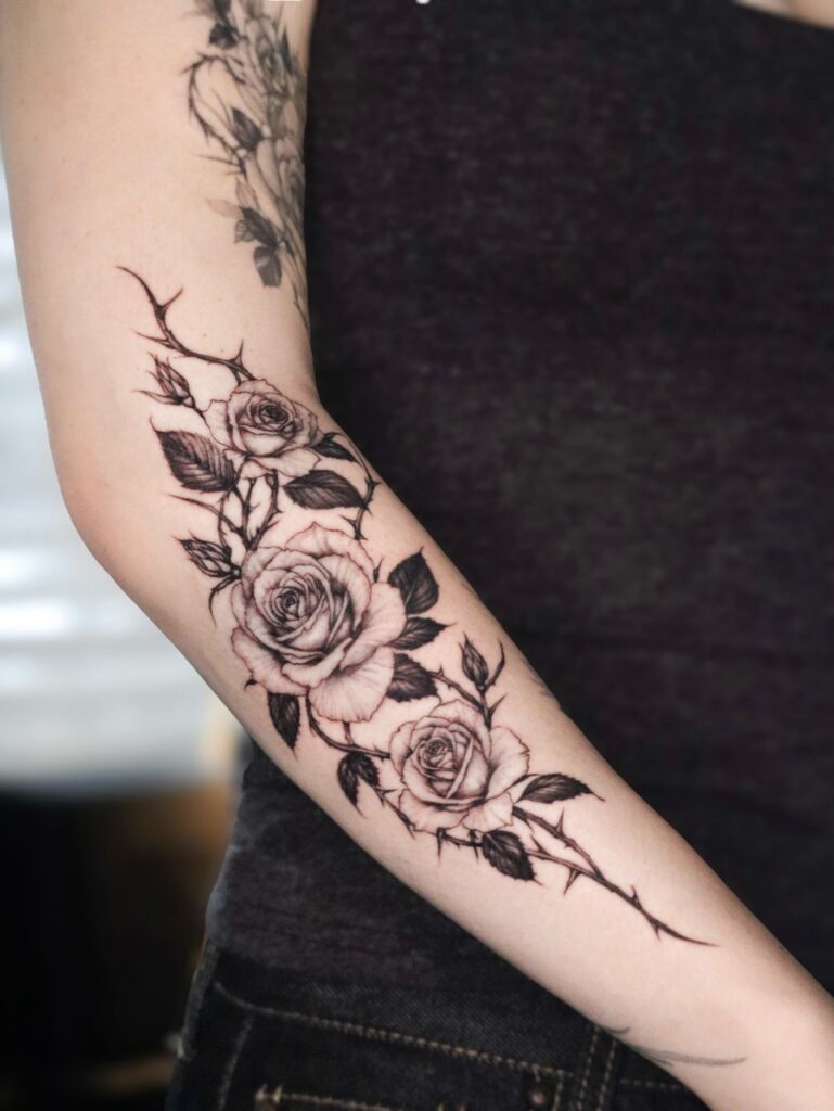 Three black ink roses tattooed vertically on a person's forearm with thorns and leaves.