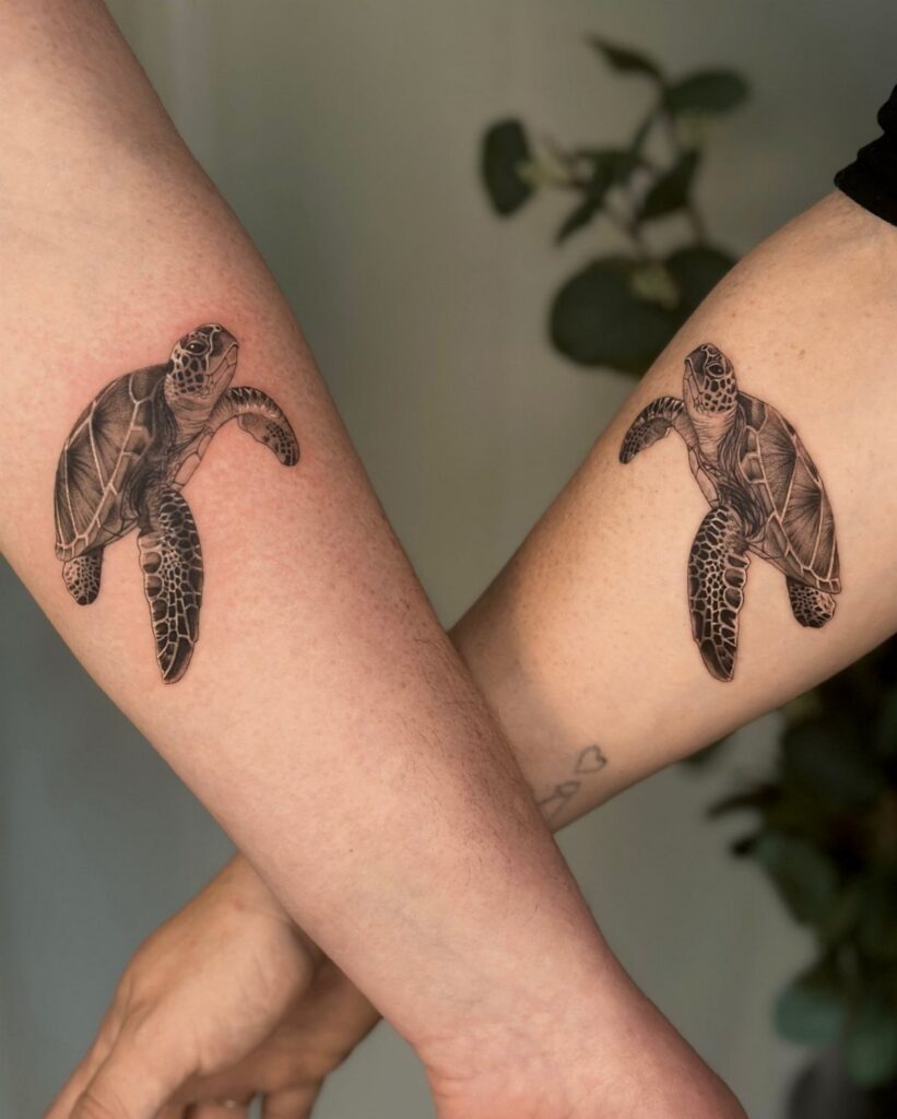 Two matching sea turtle tattoos on crossed forearms against a neutral background.