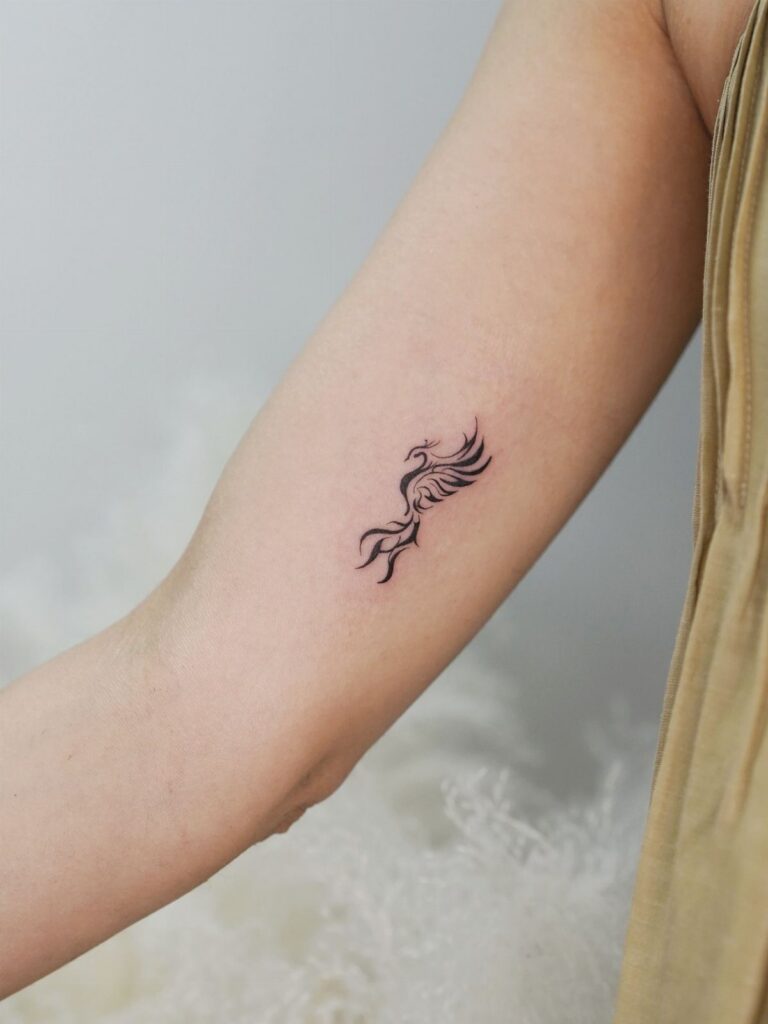 Small black phoenix tattoo on a person's inner forearm against a white background