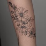 Daisy flower tattoo with leaves in black ink on a person's arm.