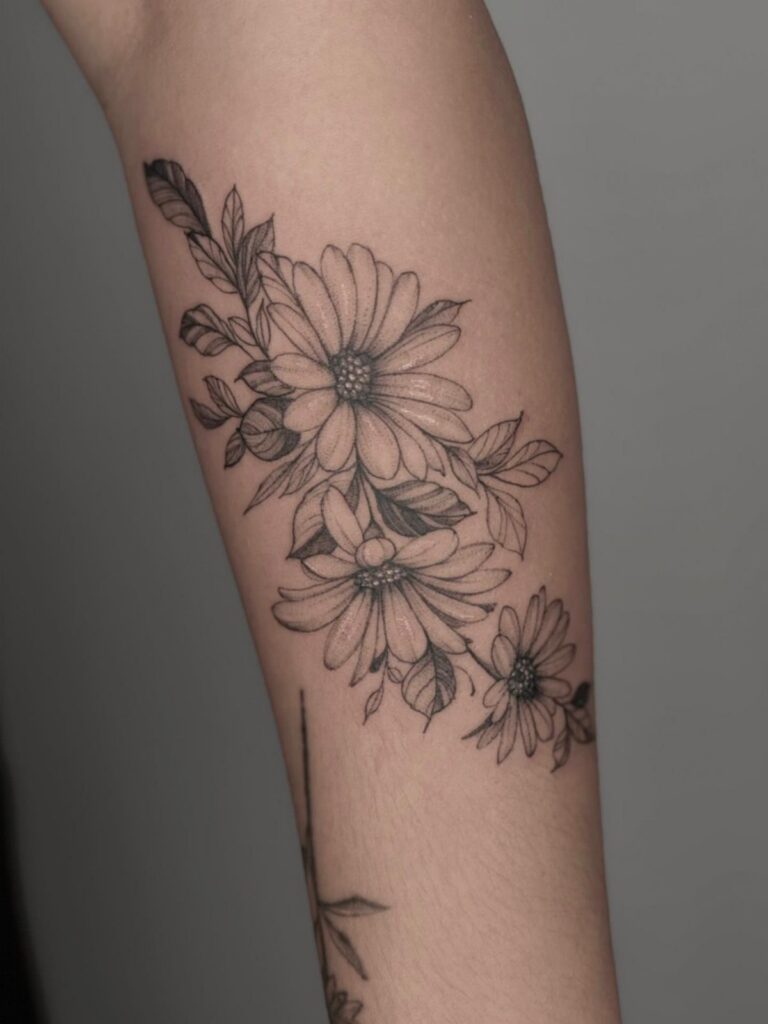 Daisy flower tattoo with leaves in black ink on a person's arm.